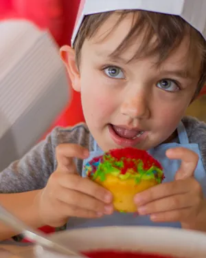 Un niño pequeño usando un gorro de chef sosteniendo felizmente un cupcake colorido y decorado.
