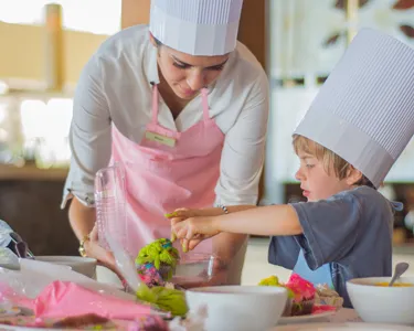 Un chef ayudando a un niño a decorar comida colorida en una mesa, ambos usando gorros de chef.