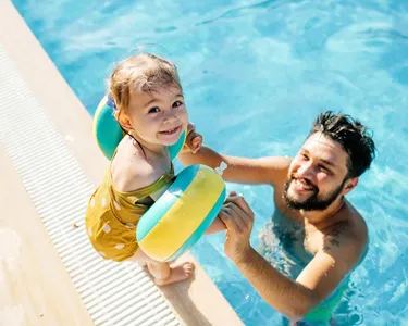 Un padre jugando con su hijo pequeño en el borde de una piscina, el niño usando flotadores en los brazos y sonriendo.