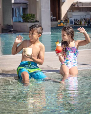 Dos niños sentados en el borde de una piscina, disfrutando bebidas coloridas mientras chapotean en el agua.