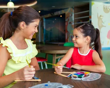 Dos niñas pequeñas sentadas en una mesa, pintando y haciendo manualidades felices en un espacio cerrado.