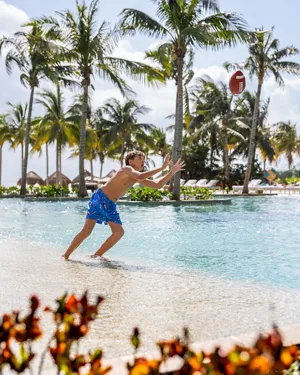 Niño corriendo al borde de la alberca intentando atrapar una pelota, con palmeras al fondo.