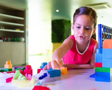 Niña concentrada construyendo una estructura colorida con bloques de plástico grandes en una sala de juegos.