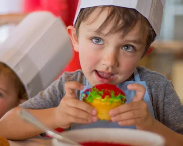 Niño pequeño con gorro de chef, emocionado mientras sostiene y prueba un cupcake decorado por él mismo.